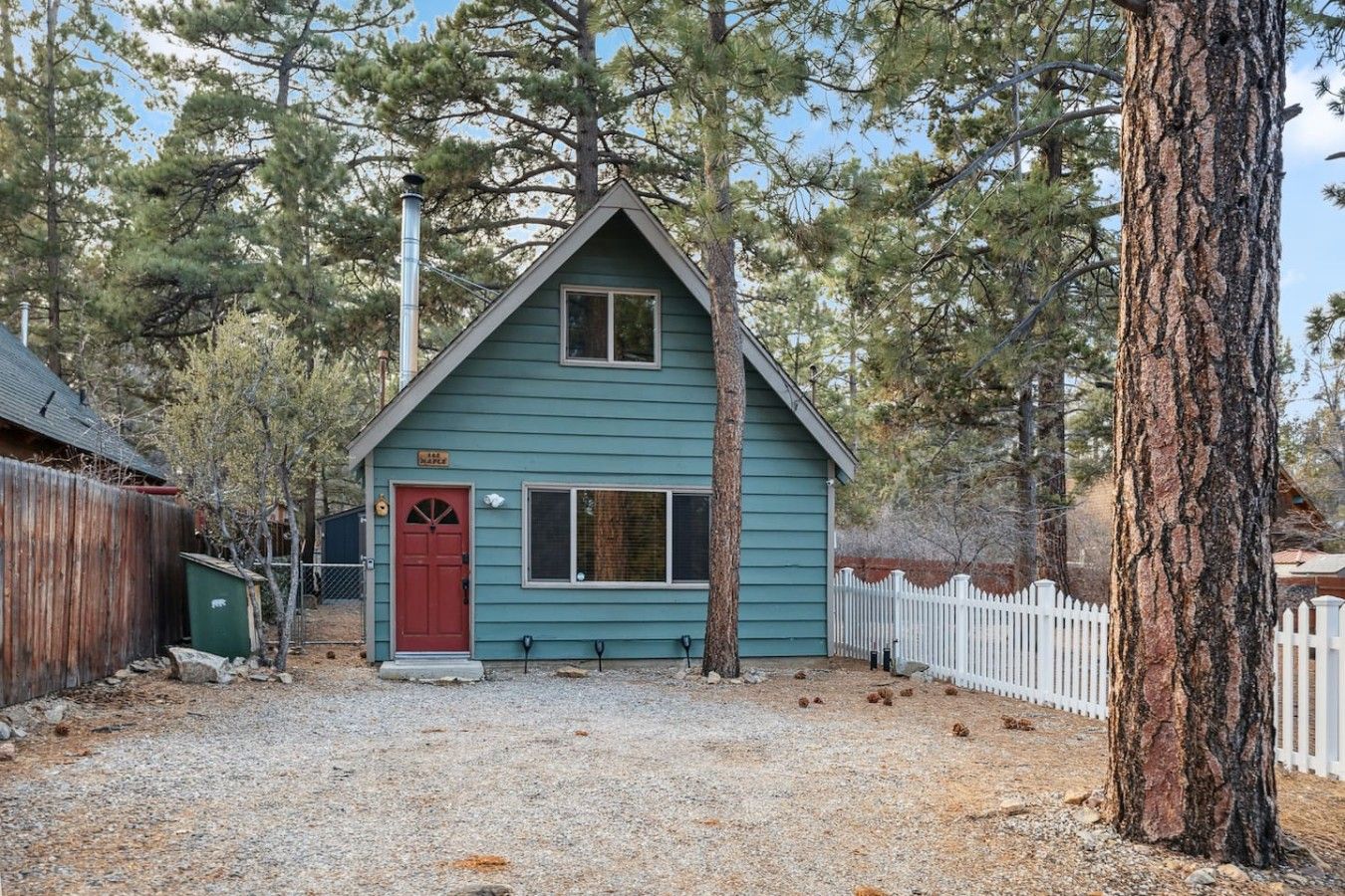 A small, single-story house with a red door and a window, surrounded by trees and a white picket fence.