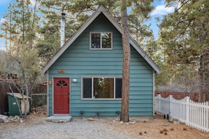 Une petite maison à un étage, avec une porte rouge et une cheminée, entourée d’arbres et d’une clôture blanche en bois.