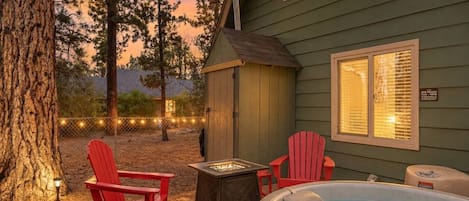 A backyard with a hot tub, two red Adirondack chairs, a fire pit, and a wooden shed.