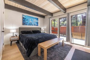 A bedroom with a large bed, a bench, a nightstand, a painting, and a sliding glass door leading to an outdoor area. - Après All Day | Walk to Summit, Community Hot Tub (Big Bear Lake)