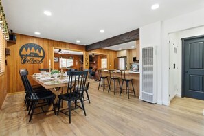 A dining area with a wooden table and chairs, a kitchen in the background, and a door on the right. - Big Bear Cabana Near Lake W/ Hot Tub, Shuffleboard (Big Bear Lake)