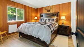 Un dormitorio con pared de madera, una cama con colcha a cuadros, dos lámparas de noche, una ventana con persianas y un radiador.