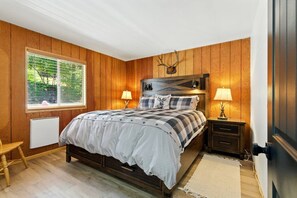 A bedroom with a wooden paneled wall, a bed with a checkered bedspread, two bedside lamps, a window with blinds, and a radiator. - Big Bear Cabana Near Lake W/ Hot Tub, Shuffleboard (Big Bear Lake)