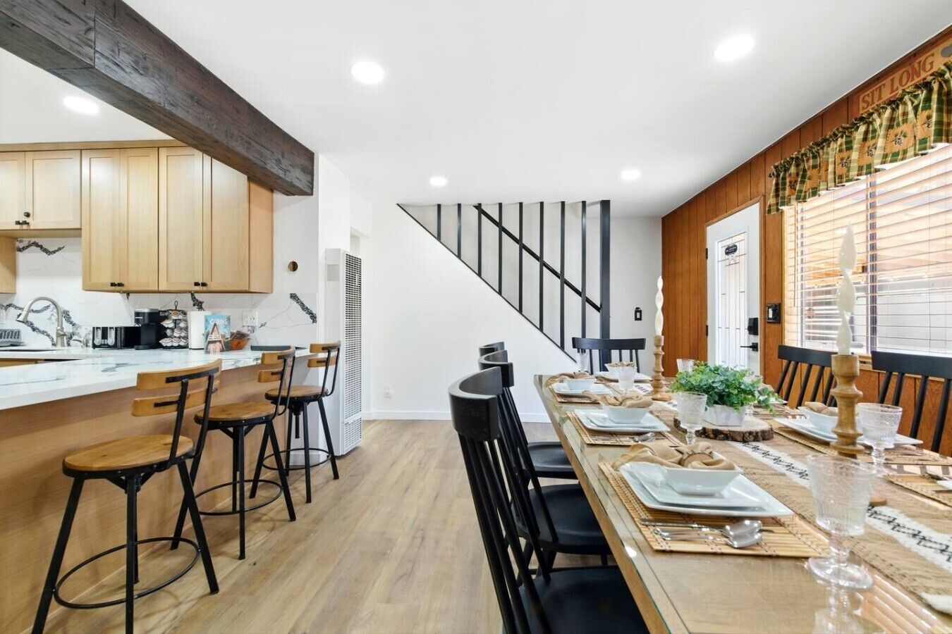A modern kitchen with wooden cabinets, a dining area with a long table set for four, and a staircase leading to another level.