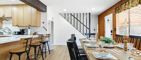 A modern kitchen with wooden cabinets, a dining area with a long table set for four, and a staircase leading to another level.