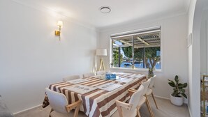 A dining area with a table set for four, a lamp, and a view of the sea through a window. - 4 Murray Avenue, Forster (Forster)