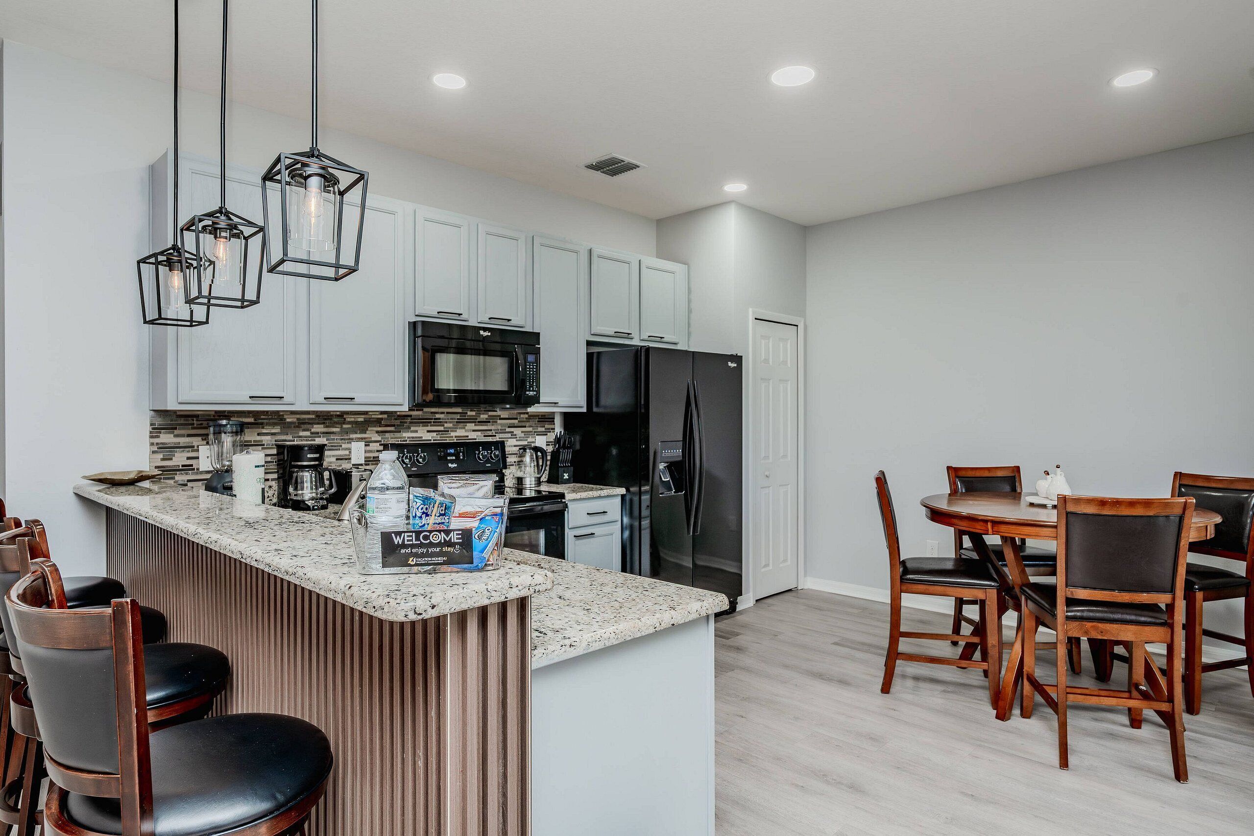 A modern kitchen with white cabinets, a black refrigerator, and a granite countertop. There is a dining area with wooden chairs and a table.