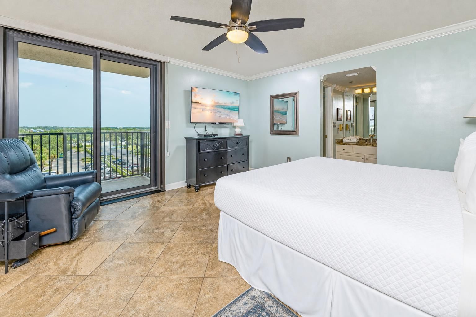 A bedroom with a large bed, a ceiling fan, a TV, and a view of the outdoors.