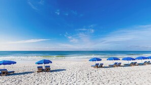 A beach with white sand and clear blue water. There are blue umbrellas and wooden lounge chairs arranged in rows. - Phoenix East 511 Gulf-Front Corner Condo (Orange Beach)