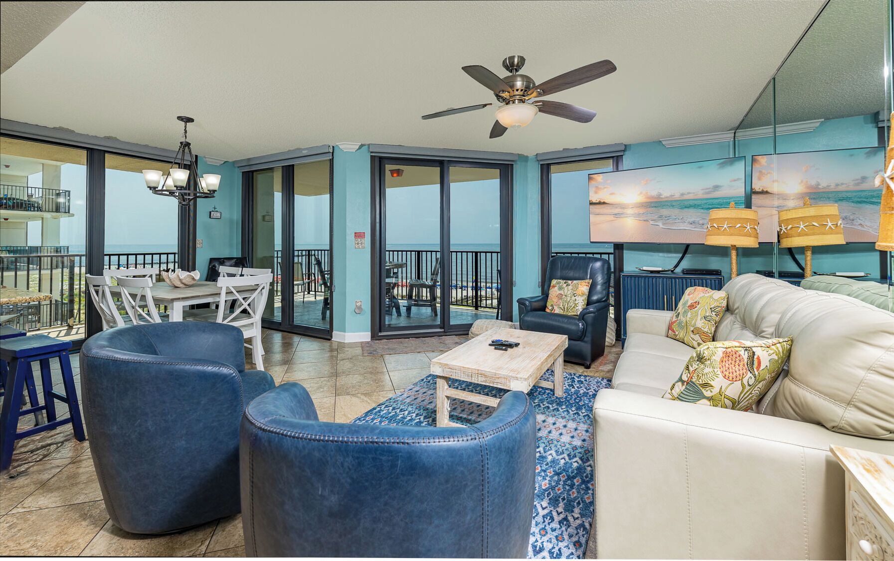 A spacious living area with a view of the beach, a ceiling fan, and a large flat-screen TV.