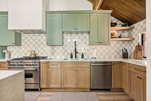 A kitchen with green cabinets, a gas stove, a dishwasher, and a checkered backsplash. - Slope & Cedar House | Hot tub, Steps 2 Snow Summit (Big Bear Lake)