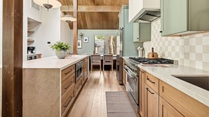 A kitchen with wooden cabinets, a central island, and a view into the dining area. - Slope & Cedar House | Hot tub, Steps 2 Snow Summit (Big Bear Lake)