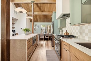 A kitchen with wooden cabinets, a central island, and a view into the dining area. - Slope & Cedar House | Hot tub, Steps 2 Snow Summit (Big Bear Lake)