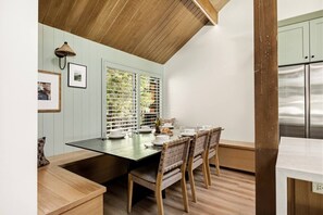 A dining area with a wooden table and chairs, a window with blinds, and a wall-mounted light fixture. - Slope & Cedar House | Hot tub, Steps 2 Snow Summit (Big Bear Lake)
