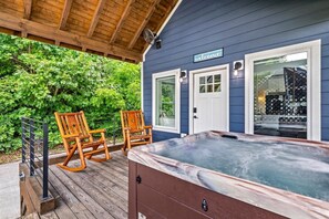 A covered outdoor area with a hot tub, two rocking chairs, and a wooden deck. - Modern Tiny Home! Walk to Parkway*Hot Tub*Sleeps 6 (Pigeon Forge)