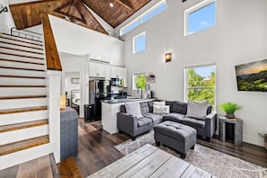 A modern interior with a staircase, a kitchen, a living area with a sofa and ottoman, and a view of greenery outside. - Modern Tiny Home! Walk to Parkway*Hot Tub*Sleeps 6 (Pigeon Forge)