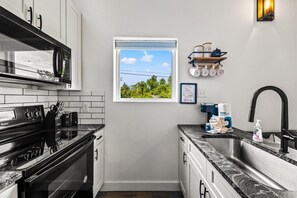 A modern kitchen with a microwave, stove, and sink, a window with a view of greenery, and a shelf with kitchenware. - Modern Tiny Home! Walk to Parkway*Hot Tub*Sleeps 6 (Pigeon Forge)