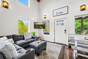 A spacious living room with a sectional sofa, ottoman, and a dining area with a bench. - Modern Tiny Home! Walk to Parkway*Hot Tub*Sleeps 6 (Pigeon Forge)