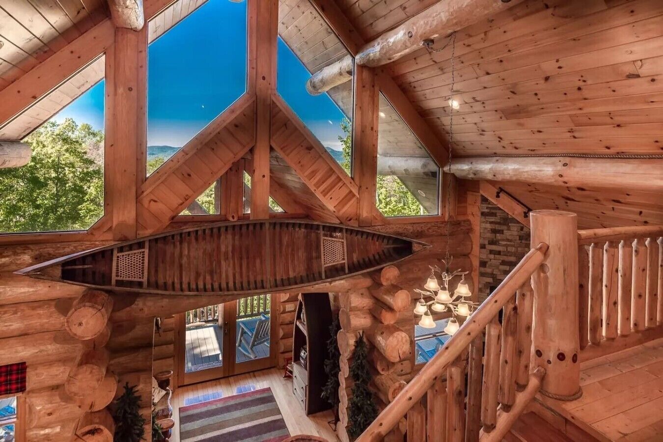 A log cabin interior with a central staircase, large windows, and a hanging light fixture.