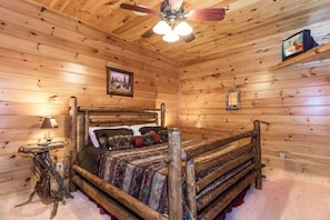 Ein Interieur einer Holzhütte mit einem Bett, einem Deckenventilator, einer Lampe und einem gerahmten Bild an der Wand.