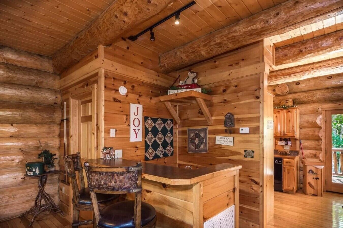 Interior de uma cabana de madeira com área de bar, uma cadeira, uma mesa e uma parede com a palavra 'JOY' e um edredom.