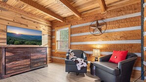 Ein Holzblockhaus-Interieur mit Holzbalkendecke, einem Fernseher links, einem Kamin und einem Sofa mit rotem Kissen.