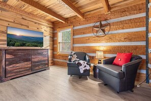 Ein Holzblockhaus-Interieur mit Holzbalkendecke, einem Fernseher links, einem Kamin und einem Sofa mit rotem Kissen.