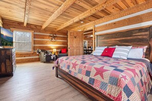 Ein Schlafzimmer in einer Blockhütte mit Holzbalkendecke, einem Bett mit einer Bettdecke mit Sternenmuster, einem Schreibtisch mit Lampe und einem Fenster mit Blick.