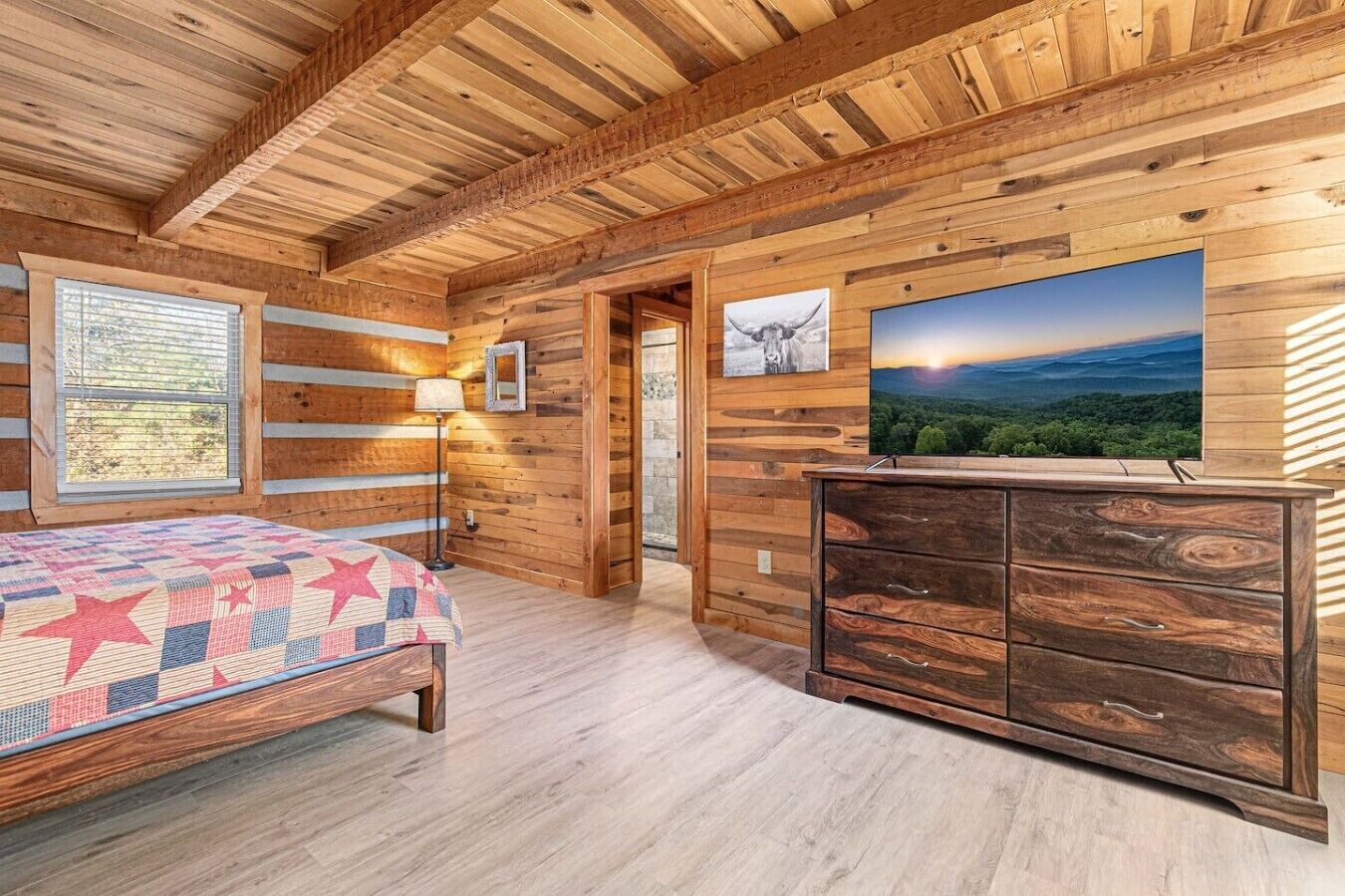 A wooden cabin interior with a bed, dresser, and a large window.