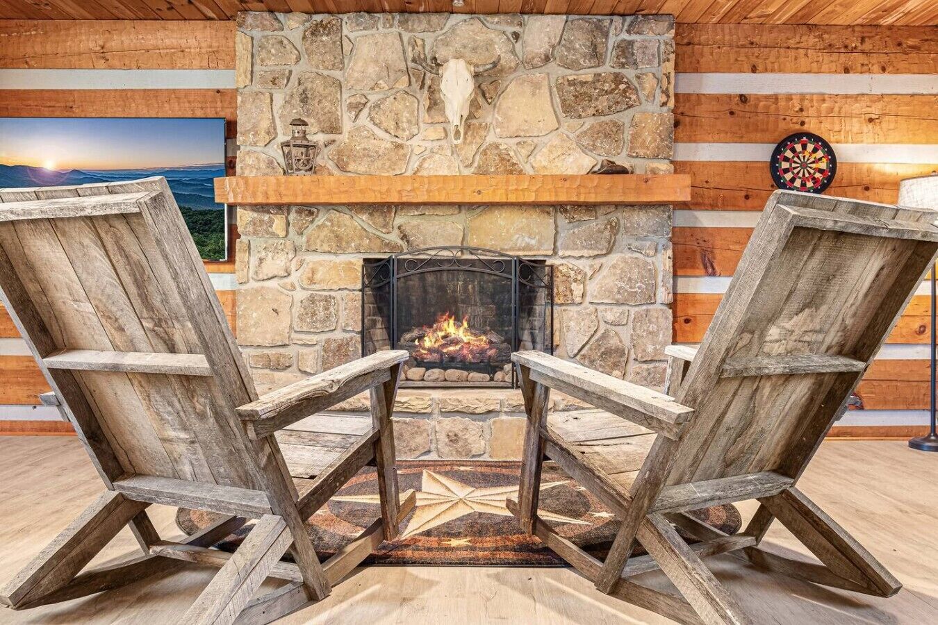 Una chimenea de piedra con dos sillas Adirondack de madera orientadas hacia ella, un dardo y un cuadro en la pared.