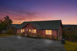 A two-story log cabin with a porch, a gravel driveway, and a view of the surrounding forest. - 5BR Pool Cabin w/MTN Views! Hot Tub*Games*Zipline! (Sevierville)