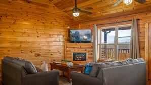 A log cabin interior with two sofas, a fireplace, a television, and a large window with a view. - 5BR Pool Cabin w/MTN Views! Hot Tub*Games*Zipline! (Sevierville)