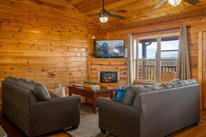 A log cabin interior with two sofas, a fireplace, a television, and a large window with a view. - 5BR Pool Cabin w/MTN Views! Hot Tub*Games*Zipline! (Sevierville)