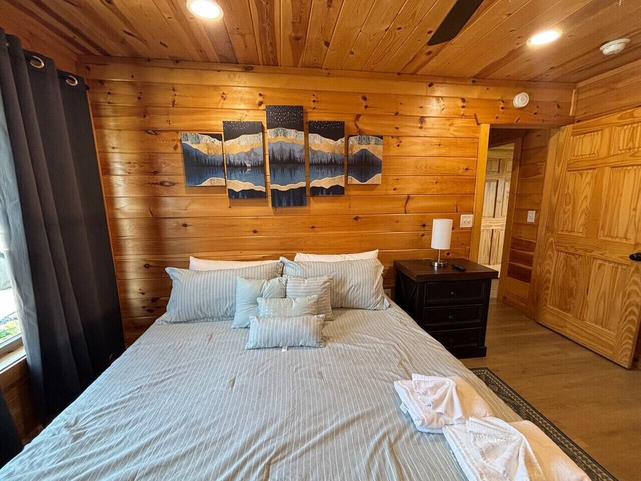 Habitación con paneles de madera, una cama, almohadas, una mesita de noche y un cuadro en la pared.