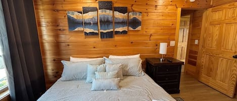 Habitación con paneles de madera, una cama, almohadas, una mesita de noche y un cuadro en la pared.