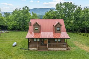 A two-story wooden house with a red metal roof, a front porch, and a grassy yard. - Creekside 3BR w/Fire Pit and Hot Tub! Peaceful! (Cosby)