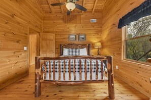 Ein Schlafzimmer einer Holzhütte mit Bett, Deckenventilator und Fenster mit Blick auf Bäume.