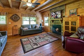 Ein gemütliches Blockhaus-Interieur mit Kamin, einer Couch, einem Ledersessel und einem Fernseher, der über dem Kamin angebracht ist.