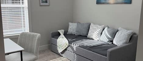 Un salon moderne avec un canapé gris, un fauteuil blanc et une petite table. Des tableaux encadrés ornent le mur, et un luminaire est fixé au plafond.