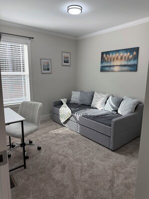 A modern living room with a grey sofa, white armchair, and a small table. There are framed pictures on the wall and a ceiling light.