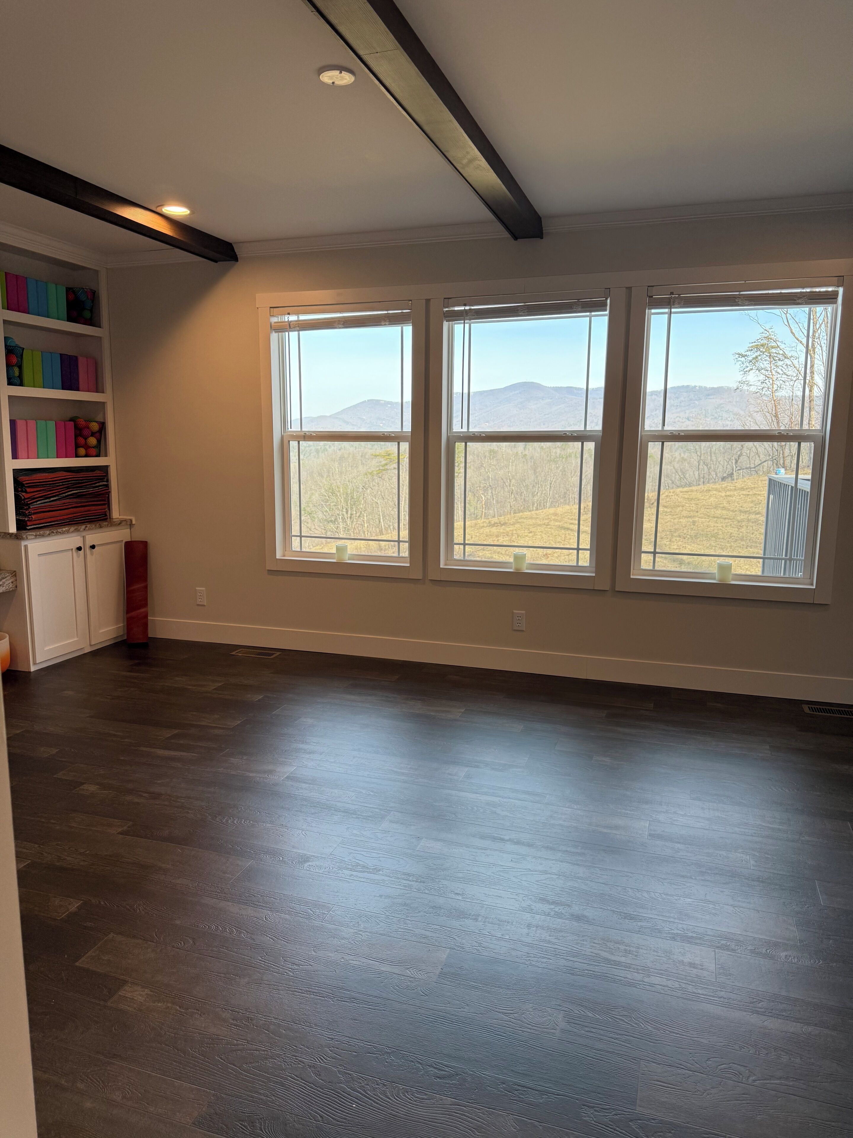A spacious room with a large window, a built-in shelf with colorful storage, and a dark wooden floor.