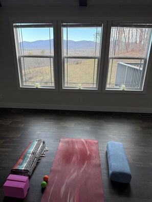 Quarto com três janelas, piso de madeira, um banco vermelho, um banquinho azul e um tapete tecido colorido.