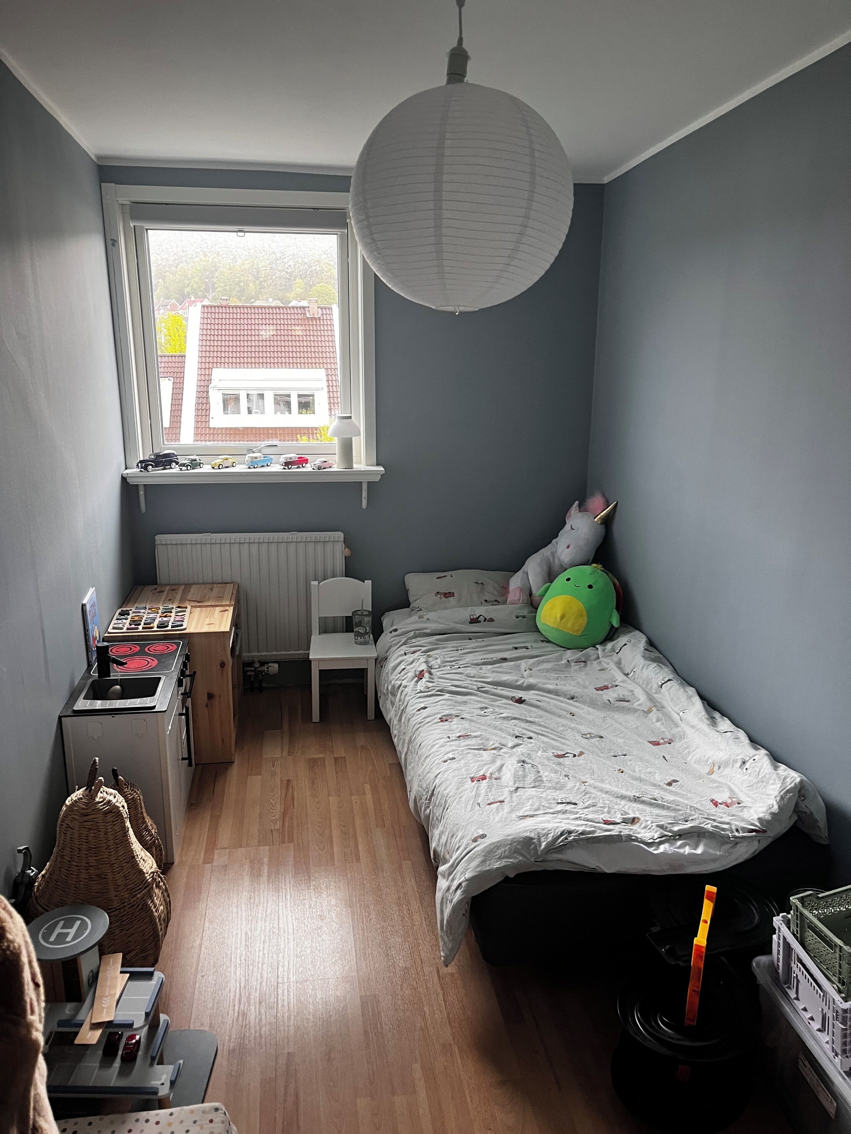 A bedroom with a bed, a window, a radiator, a toy kitchen set, and a hanging basket.