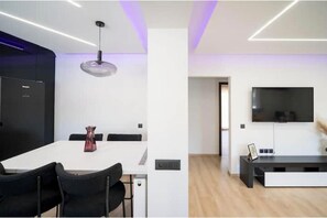 A modern living room with a flat-screen TV, a dining area with a white table and black chairs, and a pendant light. - Joy's Apartment (Chios)