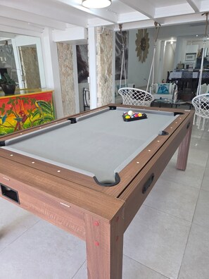 A pool table with a white cloth, surrounded by a wooden frame, in a room with tiled flooring and a dining area in the background. - Waterfront Paradise 4 bedroom Villa SXM (Koolbaai)