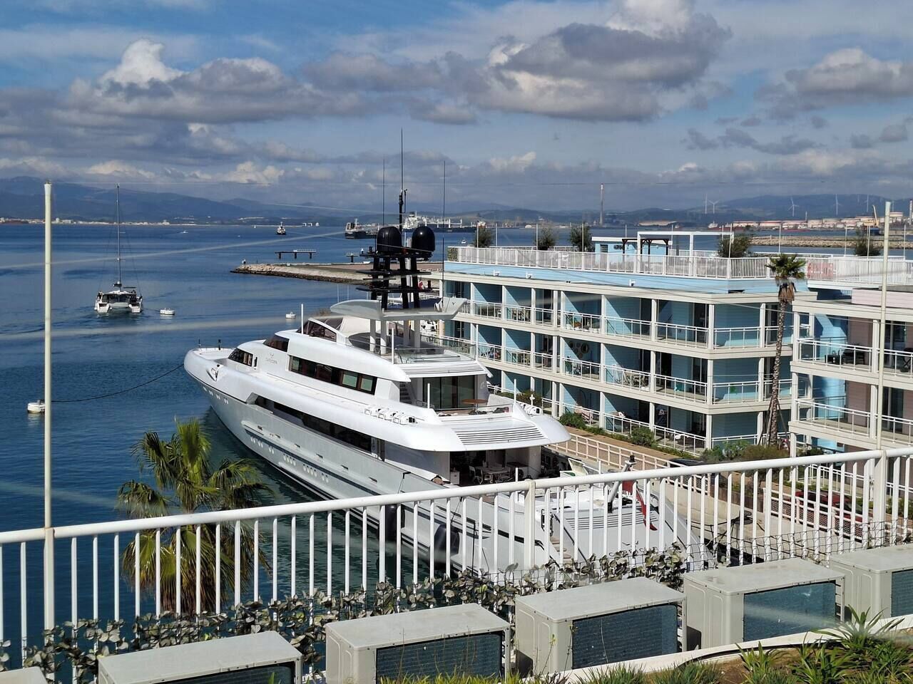 A waterfront property with a large yacht docked, a multi-story building with balconies, and a view of the sea and distant mountains.