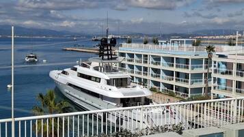 A waterfront property with a large yacht docked, a multi-story building with balconies, and a view of the sea and distant mountains.