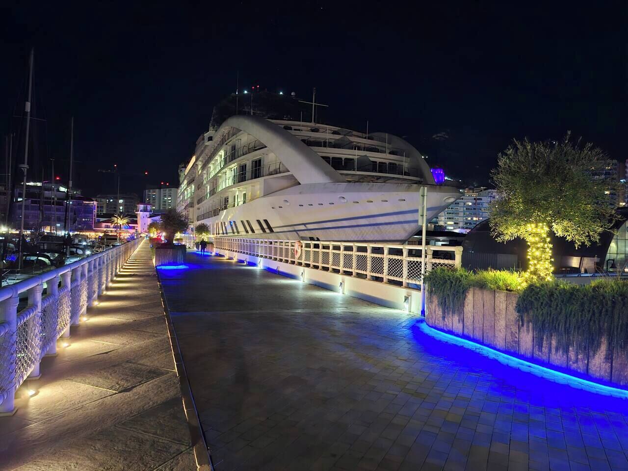 A waterfront walkway with a modern building featuring balconies and a curved roof, illuminated by blue and white lights.
