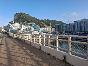 A waterfront promenade with a white fence, a cruise ship docked on the left, and buildings in the background. - Luxury Marina Resort | Rock Views & Pools - Apartment (Gibraltar)
