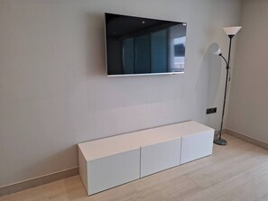A room with a mounted flat-screen TV, a white console unit, and a floor lamp. - Luxury Marina Resort | Rock Views & Pools - Apartment (Gibraltar)